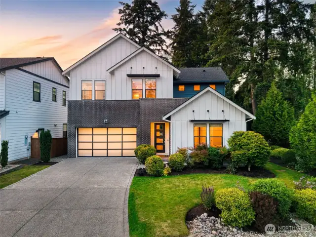 This stunning modern farmhouse blends crisp architectural lines with warm, inviting interiors, all set against a backdrop of lush evergreens. Featuring a striking brick and board-and-batten exterior, manicured landscaping, and a glowing welcome through oversized windows and a contemporary glass-panel garage.