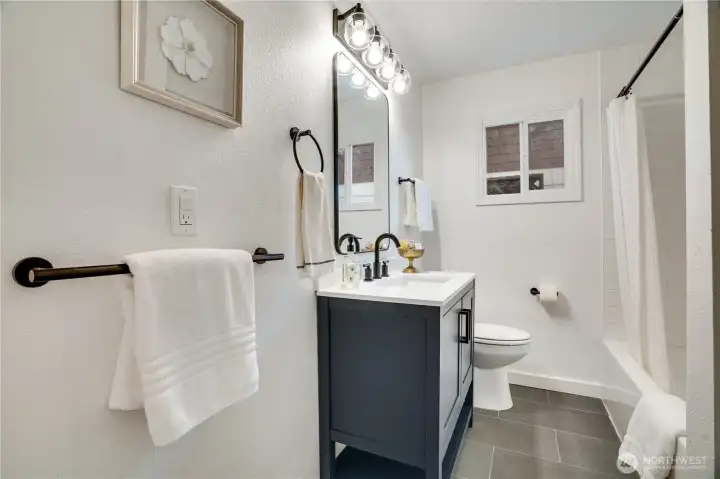 Spa-like full bath completely remodeled with a sleek charcoal vanity, matte-black fixtures, modern globe vanity lighting, white subway-tiled tub/shower combo, large window for natural light, and luxury vinyl plank flooring. Clean, contemporary, and magazine-ready.