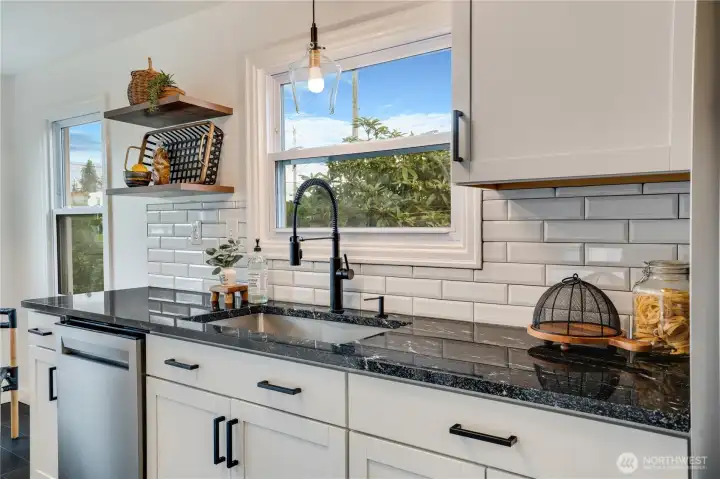 Gorgeous chef’s kitchen detail: sleek black granite counters, crisp white subway tile backsplash, matte-black spring faucet, soft-close shaker cabinets with modern black pulls, and a sunny window overlooking the backyard. Open shelving adds warmth and style. Every inch is brand new and magazine-worthy.