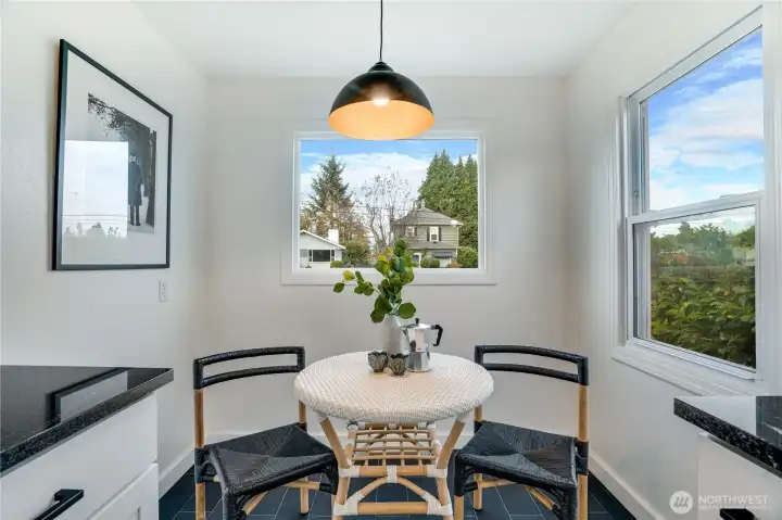 Charming built-in dining nook bathed in natural light from two large windows. Stylish black dome pendant light, sleek black tile floor, and a cozy round bistro table with woven chairs – the perfect spot for morning coffee or casual meals in this beautifully remodeled kitchen.