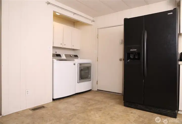 The laundry area features newer washer and dryer units that stay with the property, offering convenience and reliability. It’s easy to add a door or curtains to neatly conceal the space if desired. The door shown provides direct access to the backyard, and one of the two refrigerators is also visible in this view.