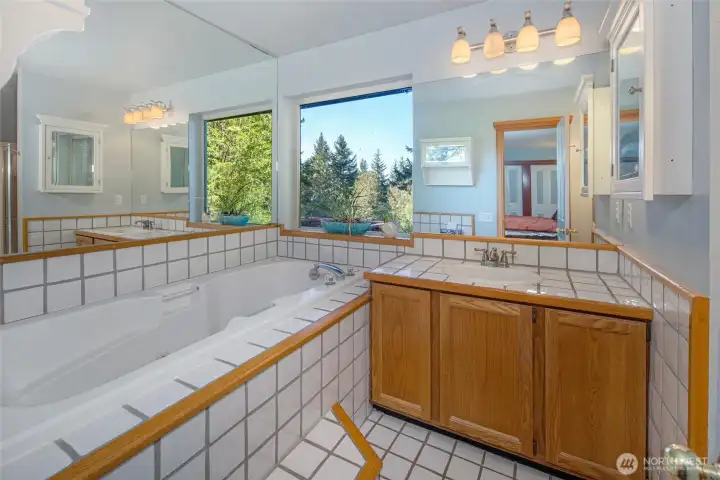 Primary bathroom with soaking tub, expansive vanity, and large picture window overlooking lush greenery—offering a spa-like setting for relaxation.