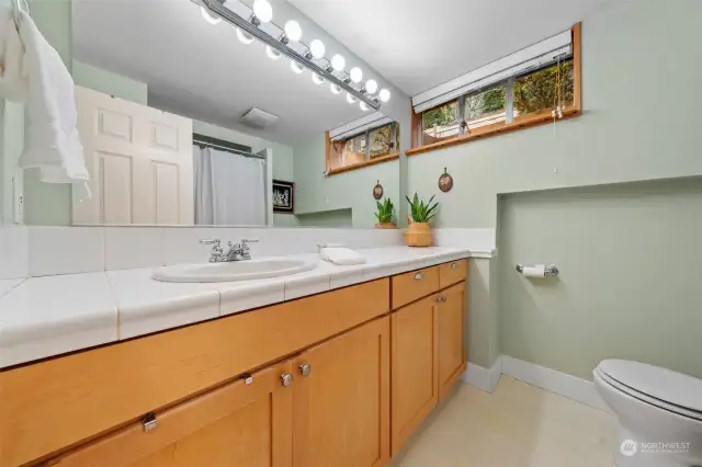 The lower-level bathroom includes a long vanity with ample storage, tiled countertops, easy-care flooring and a tile-like shower surround. Natural light from the high-set windows adds brightness and charm.