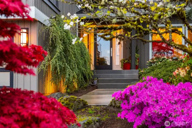 This enchanting property showcases a vibrant garden paradise with spectacular magenta azaleas in full bloom creating a dramatic focal point. A charming pathway winds through the lush landscape, leading to a cozy home with distinctive architectural character. The property features mature trees providing natural privacy and dappled shade, with splashes of yellow foliage adding warmth to the scene. Thoughtfully placed garden structures peek through the greenery, suggesting delightful outdoor living spaces. This colorful, established garden offers a magical retreat with year-round visual interest and a peaceful ambiance that welcomes you home.