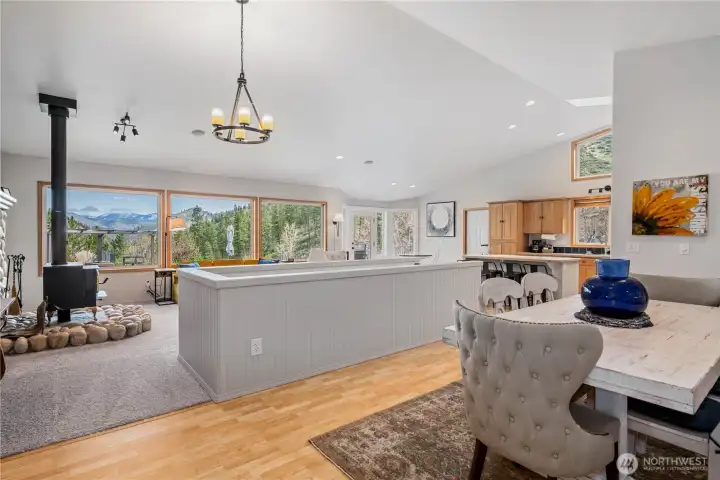 Light-filled open living with expansive windows framing mountain views, seamlessly connecting the living, dining, and kitchen spaces. Vaulted ceilings and a cozy wood stove with river rock surround create a warm yet airy gathering space.