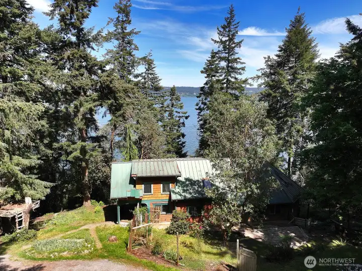Another view of the backside of the home. There is an old treehouse that could easily be brought to stability with some TLC. The upper windows are to the upper level Primary Suite. Blue sky and glistening waters!