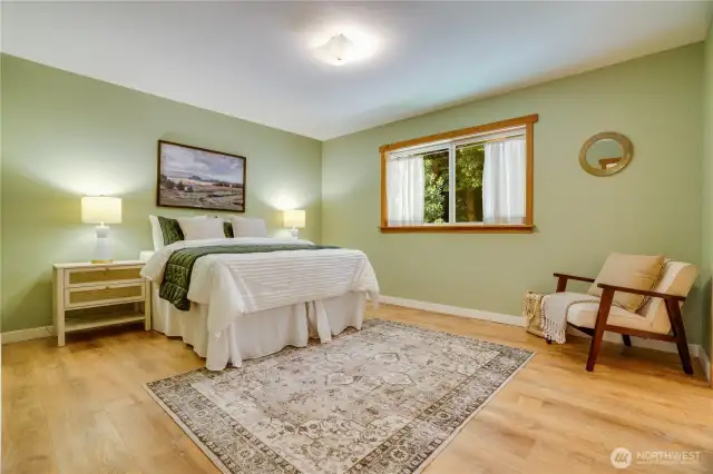 Peaceful and private primary suite on the main floor with serene green tones, wood trim, and a picture window that brings the outdoors in.