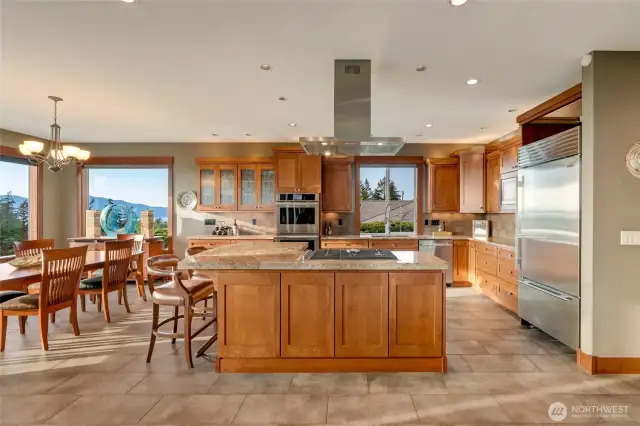 A spacious kitchen and dining area designed for both everyday living and entertaining, featuring rich wood cabinetry, a large center island, and walls of windows showcasing scenic views.
