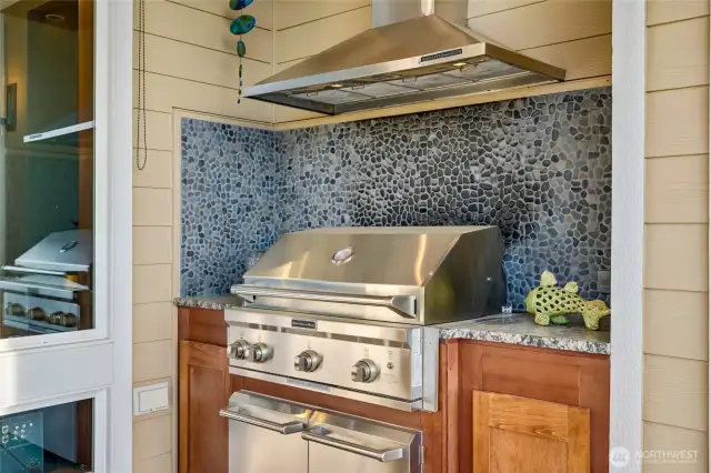The covered outdoor kitchen features a built-in stainless steel grill, granite countertops, and a stylish stone mosaic backsplash.