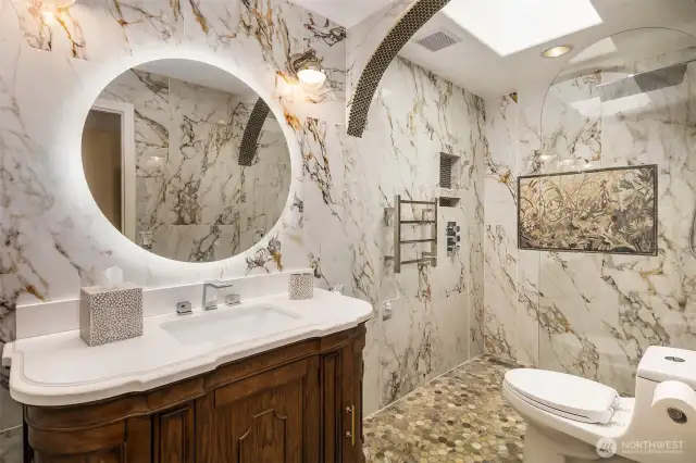 Step into opulence in this artfully designed bathroom featuring dramatic marble walls, a custom wood vanity with integrated LED mirror, a curbless walk-in shower with mosaic flooring, rainfall showerhead, towel warmer, and an elegant in-shower tapestry-style tile mural that brings timeless sophistication to every detail.