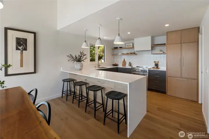 This spacious, light-filled kitchen blends luxury and function with an oversized peninsula, sleek open shelving, premium appliances, and seamless flow to the dining area and outdoor deck.