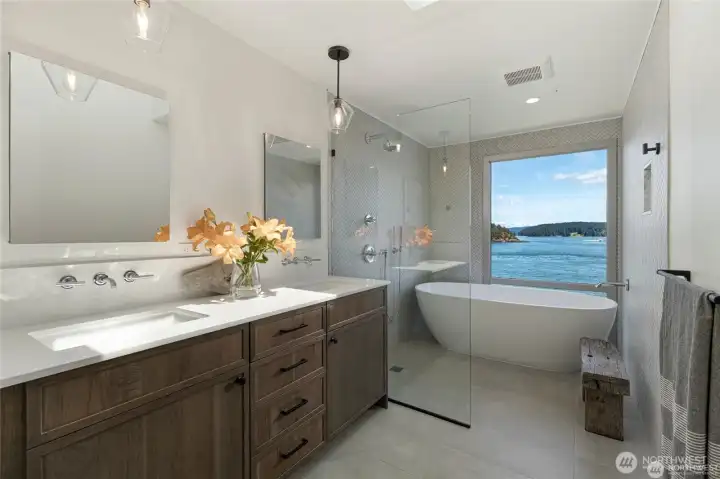 A spa-inspired primary bath showcases a freestanding soaking tub positioned before a large picture window framing blue-water vistas. The walk-in shower, elegant double vanity, and designer finishes create a luxurious retreat elevated by natural light and scenic beauty.