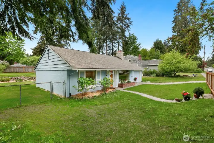 Front exterior of a mid-century ranch-style home with light blue siding, warm wood front door, brick planter, attached one-car garage, and mature evergreens on a landscaped lot.