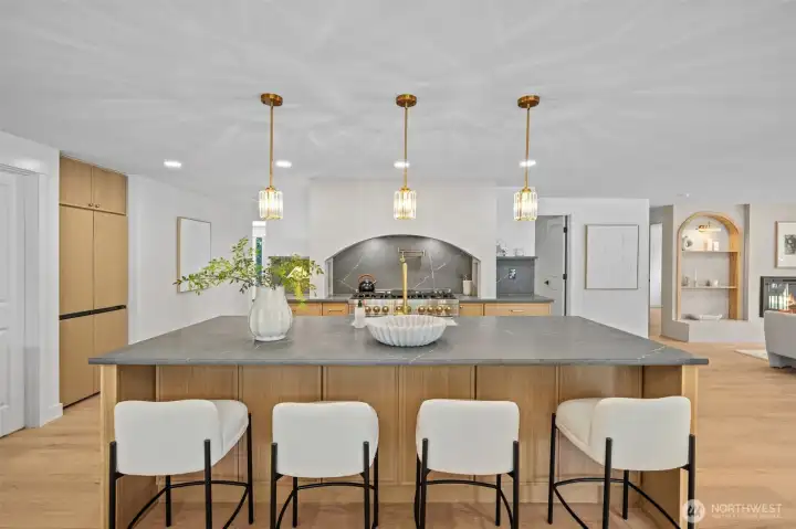 This home showcases a chef's kitchen anchored by an oversized island with honed stone countertops, illuminated by a trio of brass pendant lights. The arched plaster hood surround and slab stone backsplash frame a professional-style range with a pot filler, creating a striking focal point for the space. This home showcases a chef's kitchen anchored by an oversized island with honed stone countertops, illuminated by a trio of brass pendant lights. The arched plaster hood surround and slab stone backsplash frame a professional-style range with a pot filler, creating a striking focal point for the space.
