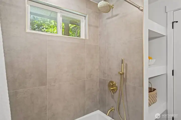 Large-format tile surrounds this shower and tub combo, complete with a rainfall showerhead, brass hand shower, and a window bringing in natural light and greenery views. Built-in shelving and matching brass fixtures add both function and polish to this shared bath. Large-format tile surrounds this shower and tub combo, complete with a rainfall showerhead, brass hand shower, and a window bringing in natural light and greenery views. Built-in shelving and matching brass fixtures add both function and polish to this shared bath.