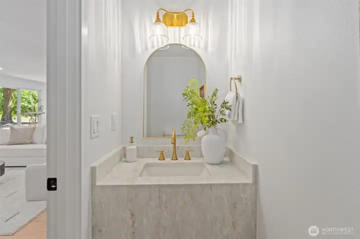 Positioned just off the great room, this powder room features a striking arched brass mirror, a dual-globe vanity light, and a full-slab stone vanity that doubles as a design statement. Brass fixtures throughout tie the space together, offering a refined touch for guests. Positioned just off the great room, this powder room features a striking arched brass mirror, a dual-globe vanity light, and a full-slab stone vanity that doubles as a design statement. Brass fixtures throughout tie the space together, offering a refined touch for guests.