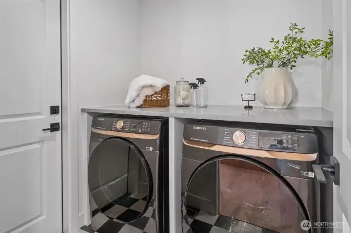 Located between the garage and kitchen, this utility room features front-load washer and dryer units set beneath a durable quartz countertop for easy folding. Bold checkered flooring adds character, while the built-in counter space makes laundry day effortless. Located between the garage and kitchen, this utility room features front-load washer and dryer units set beneath a durable quartz countertop for easy folding. Bold checkered flooring adds character, while the built-in counter space makes laundry day effortless.