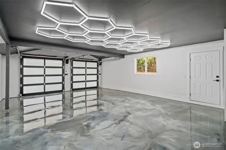 The two-car garage showcases a striking honeycomb LED lighting display overhead, paired with a glossy metallic epoxy floor and 220V wiring in place for an electric car charger. The door to the left leads you to the exterior of the home for easy access to the yard. The two-car garage showcases a striking honeycomb LED lighting display overhead, paired with a glossy metallic epoxy floor and 220V wiring in place for an electric car charger. The door to the left leads you to the exterior of the home for easy access to the yard.