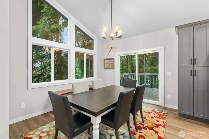 The dining area, located just off the kitchen, is framed by a wall of windows with upper panes reaching the ceiling and a glass slider that opens to the upper deck, all set against a serene, tree-lined backdrop.