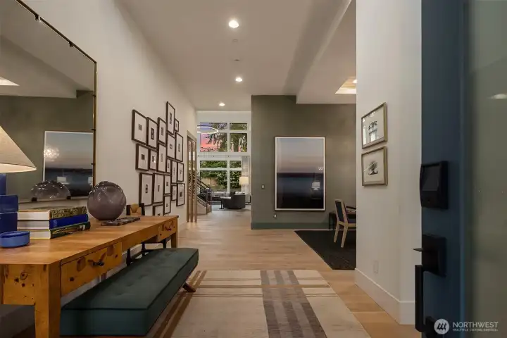A generous entry that sets the tone before you reach the great room — wide-plank engineered white oak floors, soaring ceilings, and a pink-streaked sky visible straight through to the two-story window wall beyond.
