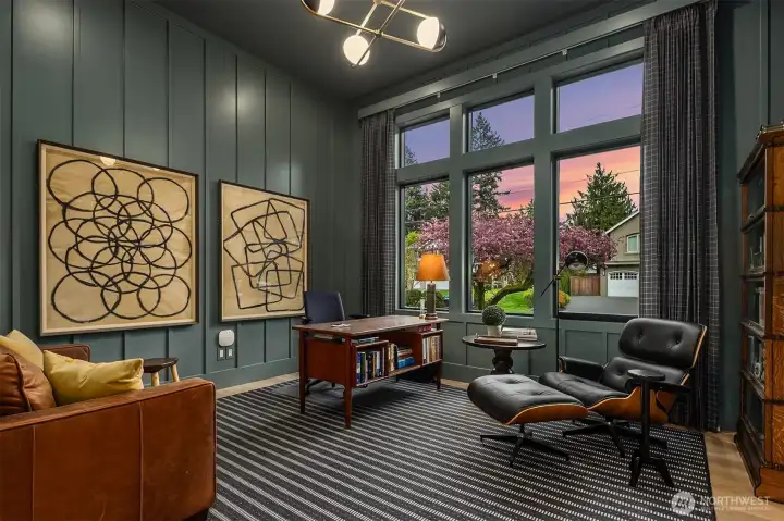 A Paquette-designed den wrapped in sage board-and-batten and custom blue plaid drapery, where floor-to-ceiling windows frame a golden-hour sky. Built for focused work during the day and too good to leave once the sun goes down.