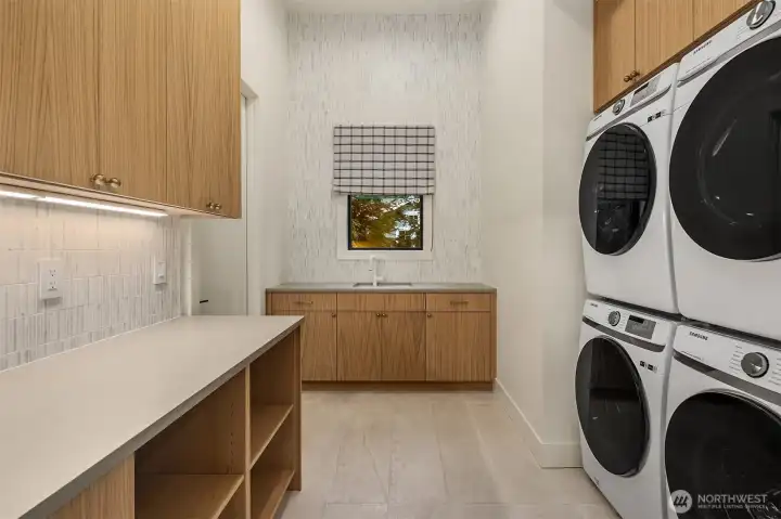 A dual-stacked laundry room that completes the primary suite with quiet efficiency — warm wood cabinetry, vertical tile backsplash, dedicated sink, and generous counter space. Even the chores feel considered here.