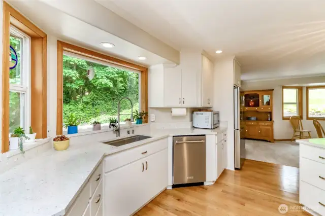 The open-concept layout flows effortlessly into a dining or living area, highlighted by gleaming hardwood floors and abundant natural light from large windows and glass doors. A large picture window above the sink provides serene views of the lush greenery outdoors, enhancing the cooking experience.