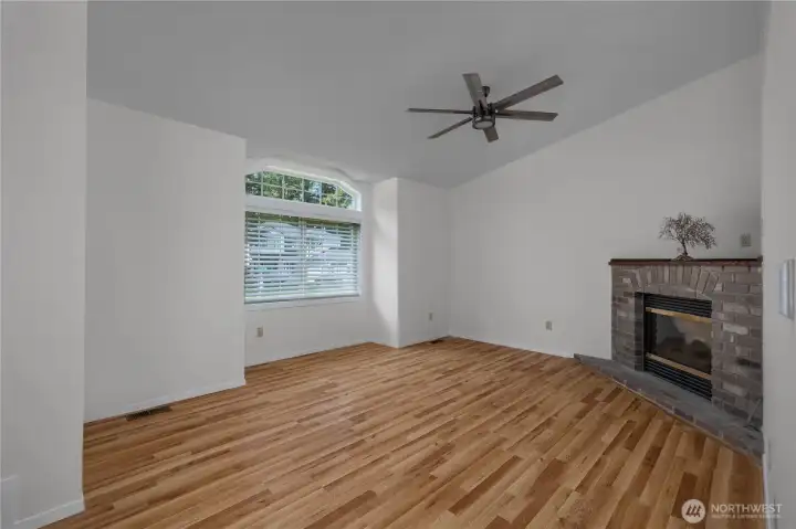 Vaulted living room with fireplace, large window, ceiling fan, and wood-style flooring.
