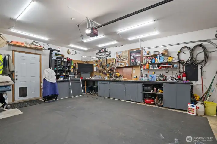 The massive garage space is a DIYer or hobbyist's dream! Outfitted with bright overhead LED strip lighting and robust rubber floor matting, it features extensive built-in lower cabinetry and sprawling workbench surfaces