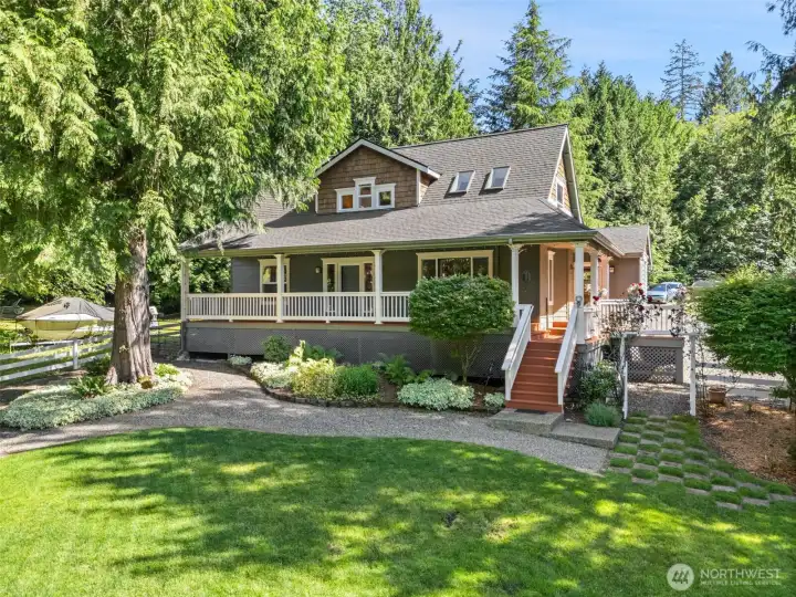 Nestled under the shade of mature evergreens, the home's expansive wraparound porch offers a quintessential Pacific Northwest vibe. Meticulously maintained garden beds with vibrant hostas and perennials frame the staircase, while the pristine white railings contrast beautifully against the warm cedar and deep grey exterior tones.