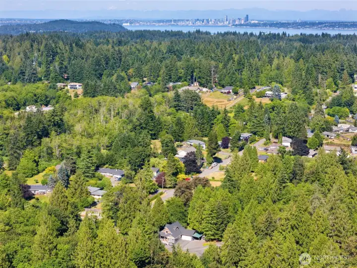 A breathtaking aerial view captures the home tucked into its lush canopy of evergreen trees, while showcasing its incredible geographic position. Looking out past the dense forest and across the shimmering waters of the Puget Sound, the iconic Seattle skyline and distant mountains sit clearly on the horizon.