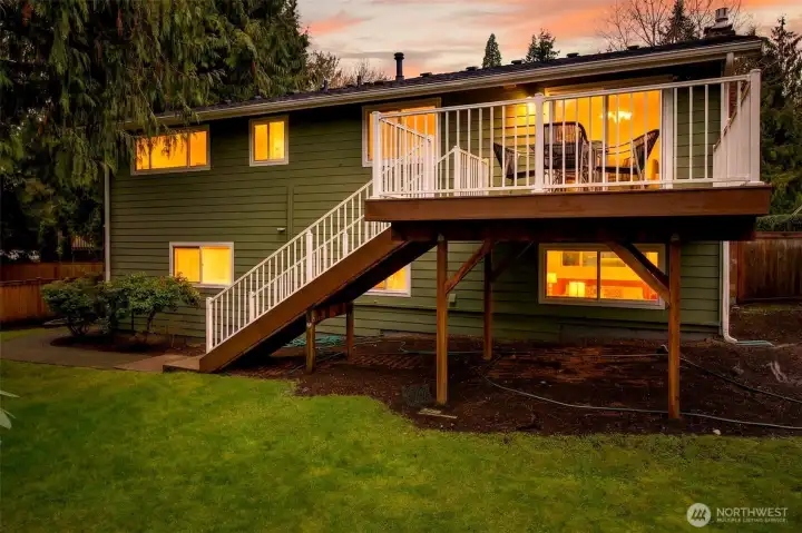 At twilight, the home takes on a warm, welcoming glow, with interior lights reflecting a sense of comfort and calm from every window. The elevated deck becomes the perfect evening retreat, framed by white railings and set against a dramatic sunset sky. This is the kind of setting that makes coming home feel special, whether you are hosting dinner or simply enjoying a quiet night in.