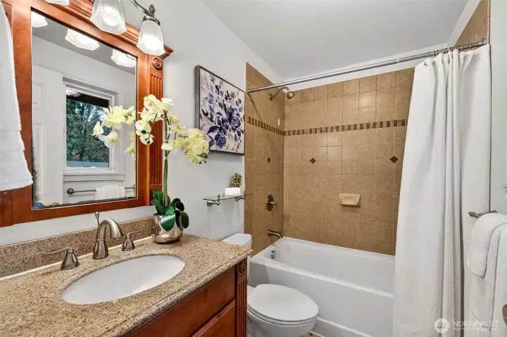 A polished bath experience with a granite vanity, an undermount sink, and updated style brushed nickel fixtures that bring a clean, finished feel. The tub and shower surround is wrapped in tile with a decorative accent band, adding character and a classic touch. A window brings in natural light, making the space feel bright and fresh.