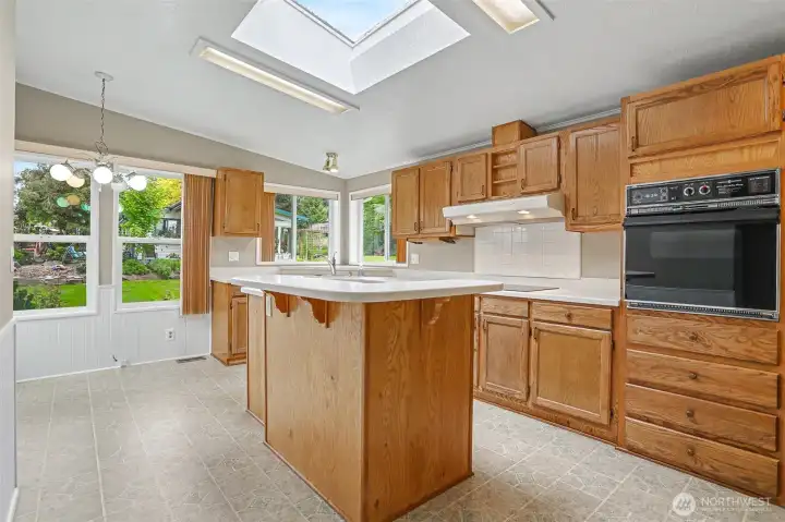 Gourmet kitchen features a center island, quartz counters, skylight, oak cabinets, wall oven, cooktop and a eating nook overlooking the community greenbelt.