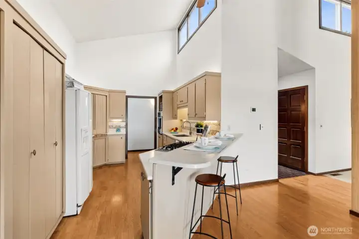 Everything about this view reinforces how well the home flows. The raised breakfast bar creates a casual place to sit or serve, depending on the moment, and it keeps the kitchen connected to the living spaces without feeling exposed. The pantry sits conveniently to the side behind folding doors, easy to access and nicely tucked away. The hardwood floors carry through seamlessly, and the vaulted ceiling with upper windows brings in a soft, natural light that keeps the whole area bright. The color combination of white walls and warm camel cabinetry gives the kitchen a grounded, welcoming feel. Look past the bar and you’ll notice a peek of the water through the family room, which adds another layer to the space. The entry and hallway toward the primary suite are also visible from here, showing how everything links together without feeling complicated.