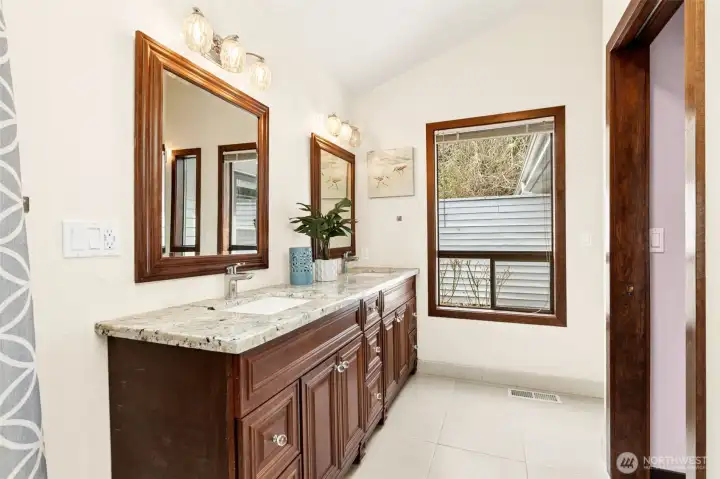 The vaulted ceiling in the primary suite bathroom gives it extra volume, and the large window brings in natural light while still keeping privacy in check with built-in blinds. The long dual-sink vanity offers plenty of space to spread out, finished with granite counters and classic cabinetry in a rich walnut tone that adds a little depth. Glass knobs give it a subtle, vintage touch without feeling dated. Tile flooring keeps everything practical and low maintenance, which is exactly what you want in a space that gets daily use.