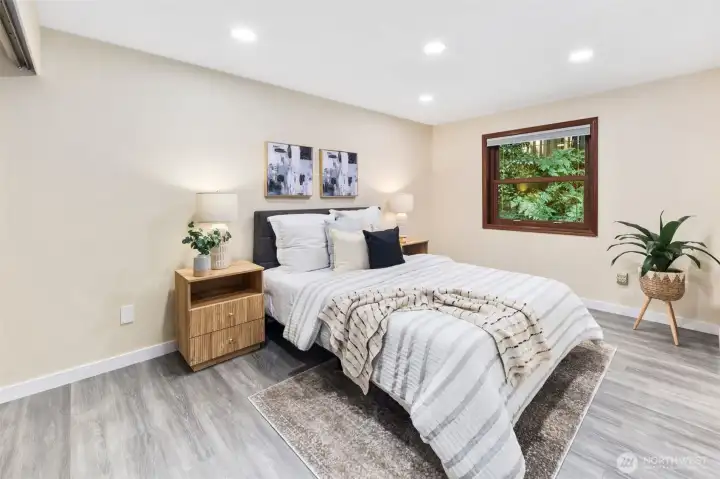 This main floor bedrooms offers comfort and convenience, with recessed lighting, warm wall tones, and a window framed in wood casing that looks out to the lush greenery of the backyard.