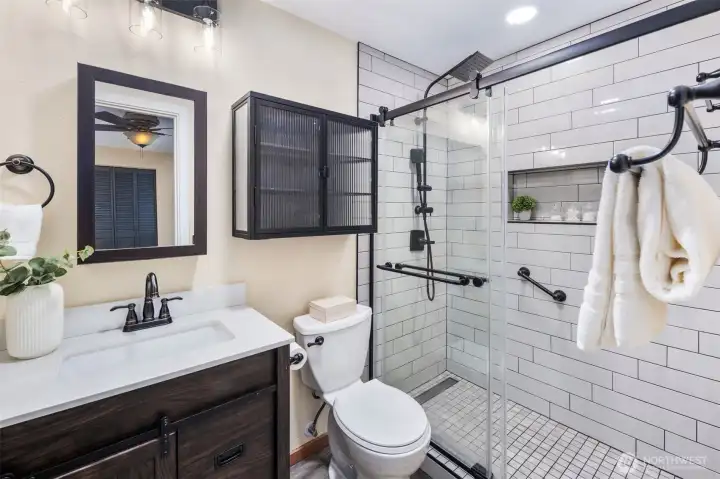 The primary bath was fully renovated in 2024 with a new vanity, sink, toilet, and fixtures, all in a sleek matte black finish to match the hardware throughout the home. Subway tile with niche shelving complements the frameless glass shower doors and modern rainfall showerhead. A three-light vanity fixture with clear glass shades adds a touch of modern industrial style, echoed by the wall-mounted storage cabinet with ribbed glass doors. Recessed ceiling lighting completes this bright, spa-like retreat.
