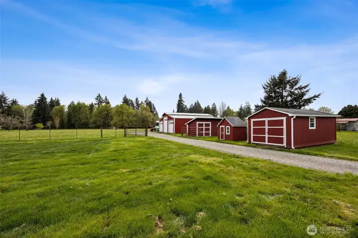 The newer gravel driveway carries you past a lineup of outbuildings that feel both practical and full of personality, with fenced and gated pasture areas unfolding around them. First up is the pig house, updated with new siding and flooring, and still sitting on its original steel skids, a movable little piece of property history. Next to it, the former chicken coop got a second act as a kids playhouse, which is just plain charming. Continue down and you reach the garden shed with new flooring and dual doors, formerly serving as a goat shed. Then comes the larger three-bay barn and shop, bringing serious utility to the mix. And just beyond, out of frame, even more infrastructure with a doghouse and feeding shed. It is hard not to appreciate how much has already been created here. This is not raw acreage waiting for years of work. It feels established and ready to be enjoyed.