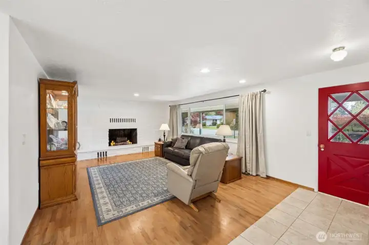 That red front door makes a statement before you even step inside, and the diagonal lattice glass panel gives it that classic mid-century farmhouse character that feels collected rather than trendy. The tile entry is a very smart landing zone too, ready for muddy boots, paws, and all the realities acreage living can bring, while protecting those gorgeous oak hardwood floors stretching through the living room. And then the fireplace takes over. An entire wall of vintage mid-century stone, painted white to blend beautifully with the bright interior, creates the kind of focal point newer homes struggle to replicate. Add the huge picture window pouring in natural light and this room lands as warm, substantial and full of character.
