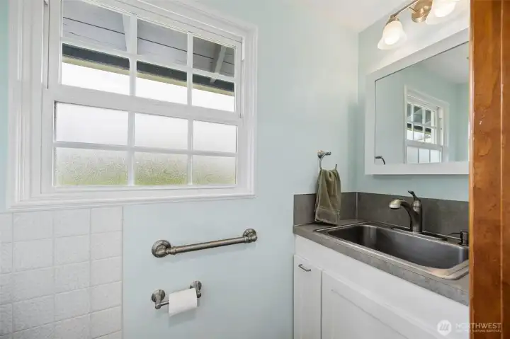 Tucked just off the kitchen near the laundry, this half bathroom has a hardworking utility side thanks to that deep stainless sink, perfect for cleanup, projects, or the kinds of tasks acreage living occasionally brings indoors, but it also happens to be genuinely charming. The whisper-blue paint gives the room a soft, calming tone, paired with crisp white tile and white shaker cabinetry that keep it feeling fresh and timeless. Luxury vinyl flooring carries through for durability, and the privacy glass on the lower half of the window lets in beautiful natural light while keeping things discreet.