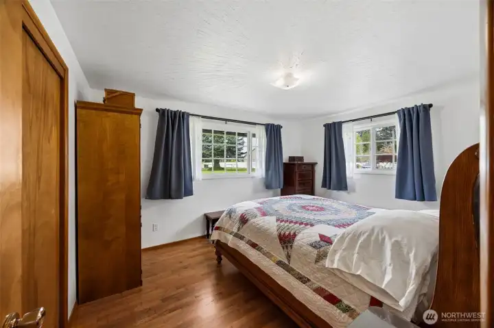 Two windows bring in sunlight from multiple directions into this primary bedroom, doubling the brightness and giving the room a wonderfully open feel, while those oak hardwood floors carry through with the same warmth found throughout the home. Fresh white walls keep it all crisp and ready for whatever style comes next. And then there is a detail you do not see every day, a subtle original ceiling texture in a starburst pattern radiating gently from the central light, almost like soft sun rays. It is understated, but incredibly charming, the kind of mid-century character people try to recreate and rarely do. No popcorn here, just a little architectural personality quietly hiding overhead.
