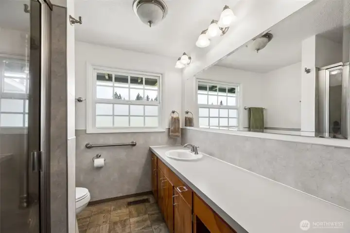 This bathroom has the sort of vintage character people try to recreate. That extra long vanity and seemingly endless counter space feel almost indulgent, paired with extensive cabinetry and an oversized beveled mirror that amplifies the whole room. There is a wonderful mid-century sensibility here, but with practical updates where they count. The window may be the cleverest detail of all, privacy glass below for comfort, clear glass above so you still catch sky and treetop views while natural light pours in. It changes the feel of the room. Waterproof vinyl flooring handles real life beautifully, and the walk-in shower keeps things streamlined and functional.