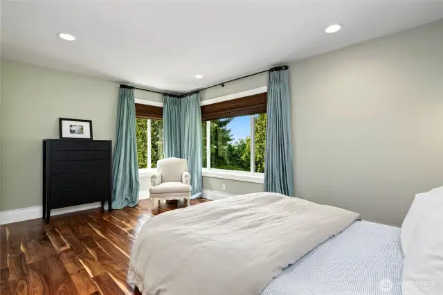 Sunlit second lower level bedroom retreat with rich hardwood floors, peaceful green-toned walls, and corner windows offering treetop views and tranquil natural light.
