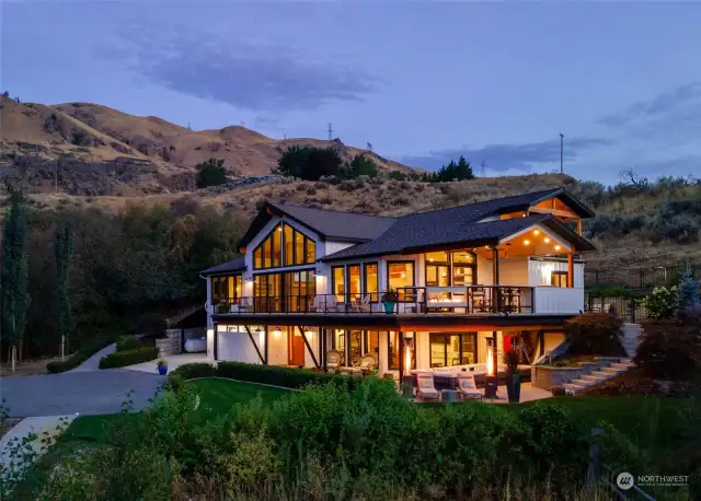 Twilight exterior view of home w/ attached garage, main level view deck w/ outdoor kitchen, dining area and lower level patio w/ hot tub/swim spa and seating area and surrounding landscape set on 12 acres.