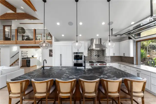 View of Main Level Kitchen w/ automated roll up garage door pass through, leather finish granite island w/ seating, high-end appliances, tile backsplash and glass sliding doors leading to the wraparound deck with outdoor kitchen, dining and lounge areas.