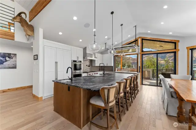 View of Main Level Kitchen w/ automated roll up garage door pass through, leather finish granite island w/ seating, high-end appliances, tile backsplash, dining room and glass sliding doors leading to the wraparound deck with outdoor kitchen, dining and lounge areas.