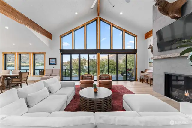 Main Level Living Room w/ high ceilings, grand fireplace and glass sliding glass doors leading to the wraparound deck w/ outdoor kitchen, dining and lounge areas.