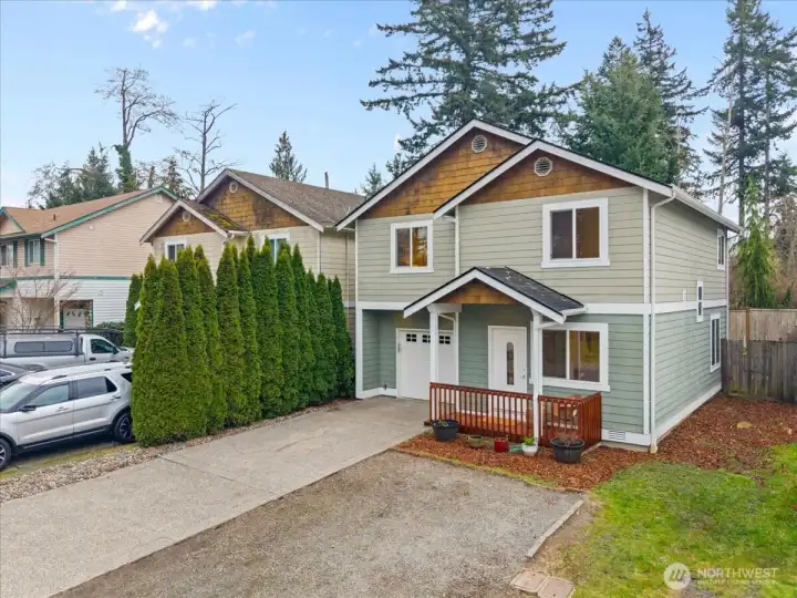 Angled exterior view highlighting the home’s gabled rooflines, covered porch, and attached garage. Tall evergreen hedges along the side add greenery and privacy.