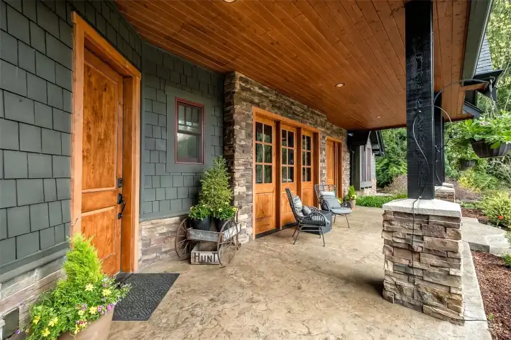 The expansive covered front porch offers year-round enjoyment with its warm wood-paneled ceiling, handsome stone accents, and a row of glass-paneled wood doors that bring in natural light while maintaining a cozy, sheltered feel—perfect for morning coffee or evening unwinding.
